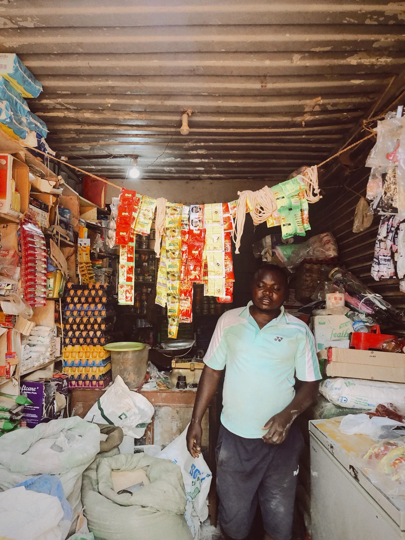 A shop owner in his crowded provision shop, goods hanging from the ceiling and stacked to the roof
