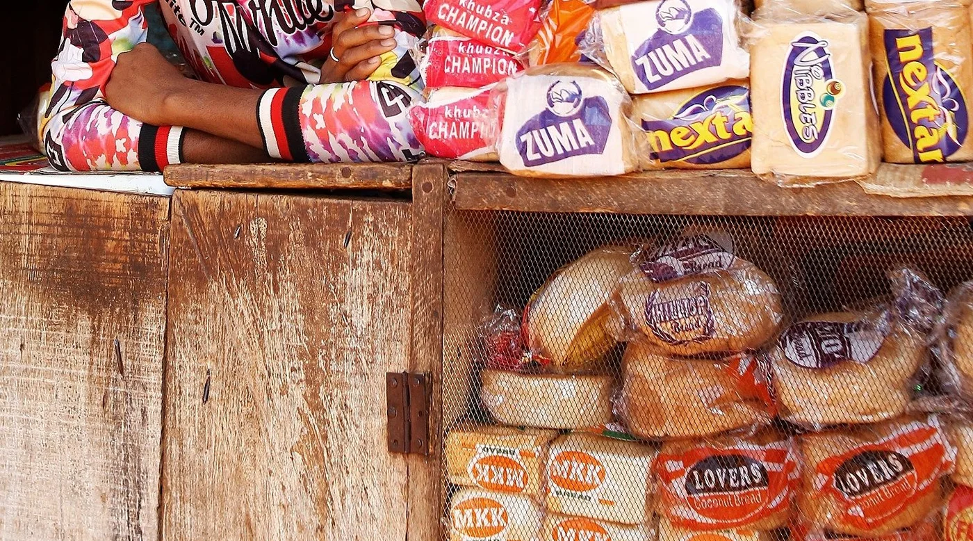 A shop attendant's folded arms on a kiosk counter, beside stacks of bread and goods