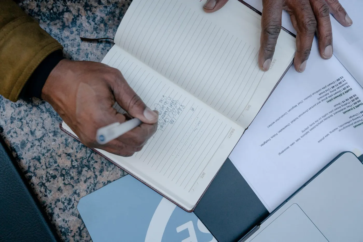 A hand writing records in a notebook among loose papers